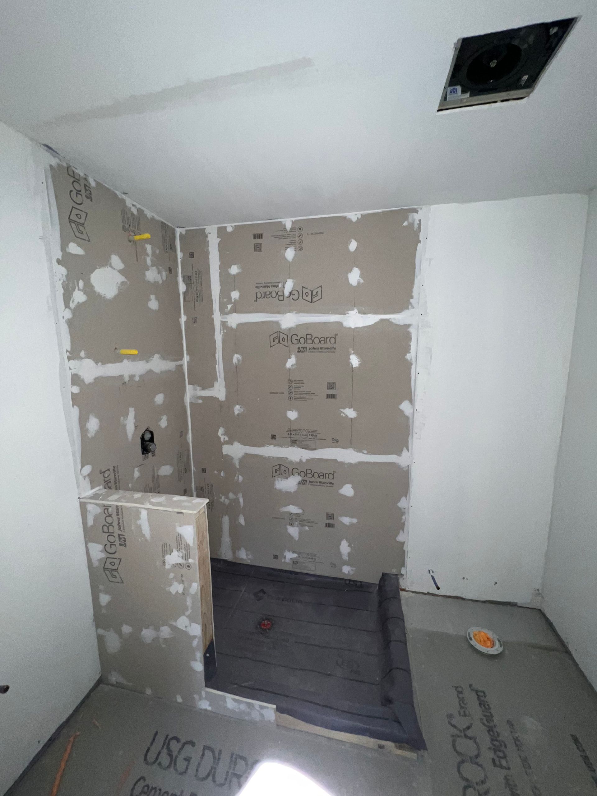 Bathroom under construction with gray cement board walls and floor. Black shower base. Ceiling with vent.