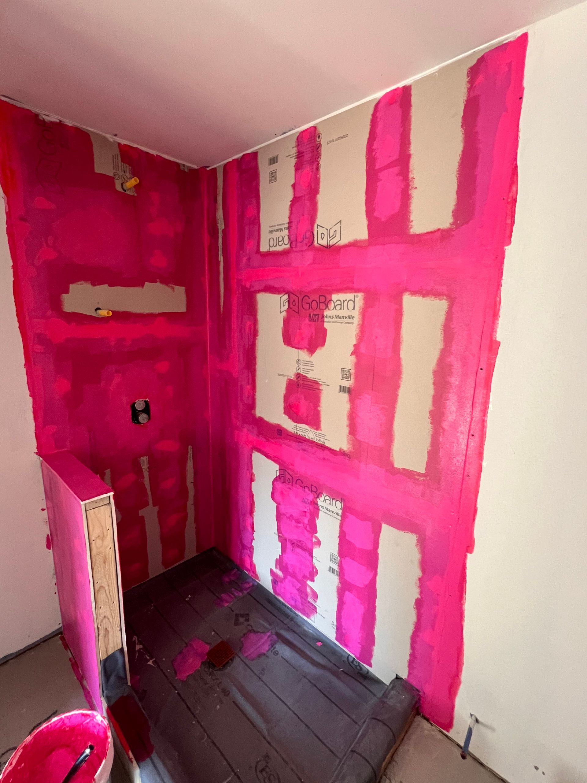 Shower stall walls covered in bright pink waterproof membrane, with exposed cardboard patches.