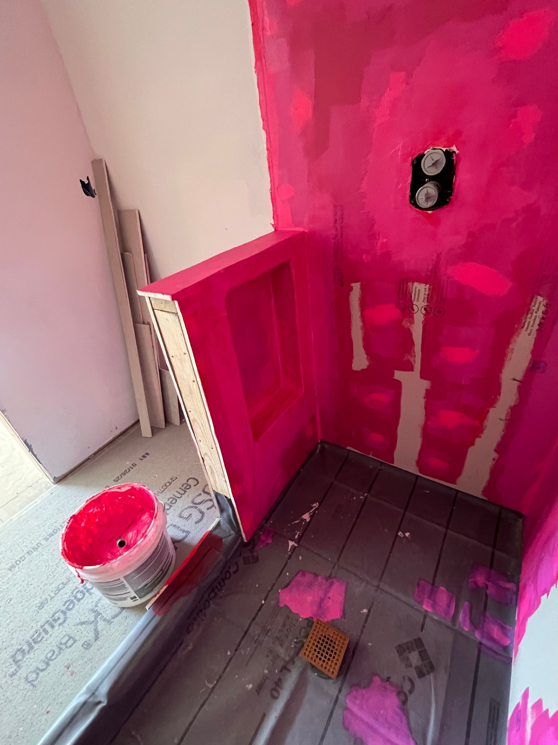 Pink-painted shower stall under construction; gray floor, white drywall, red paint bucket, outlet.