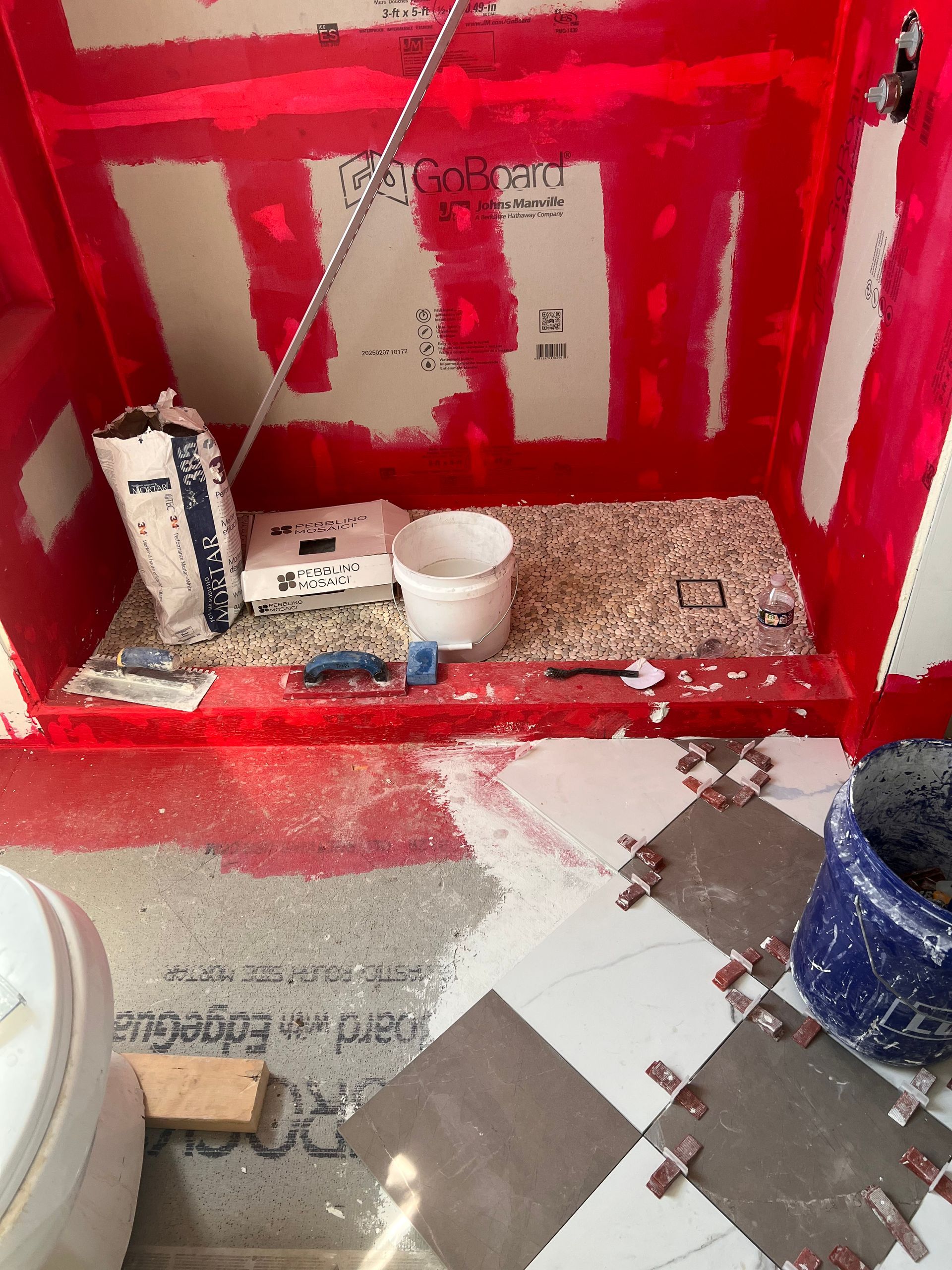 Shower construction in progress: red waterproofing, materials, and floor tiles.