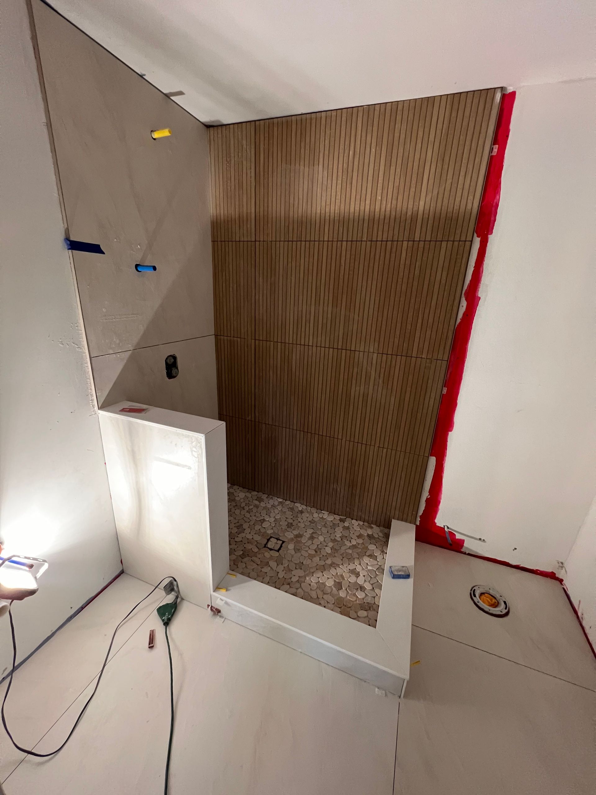 Bathroom shower under construction; beige and wood-toned walls. Tiled floor. Tools and electrical cords.