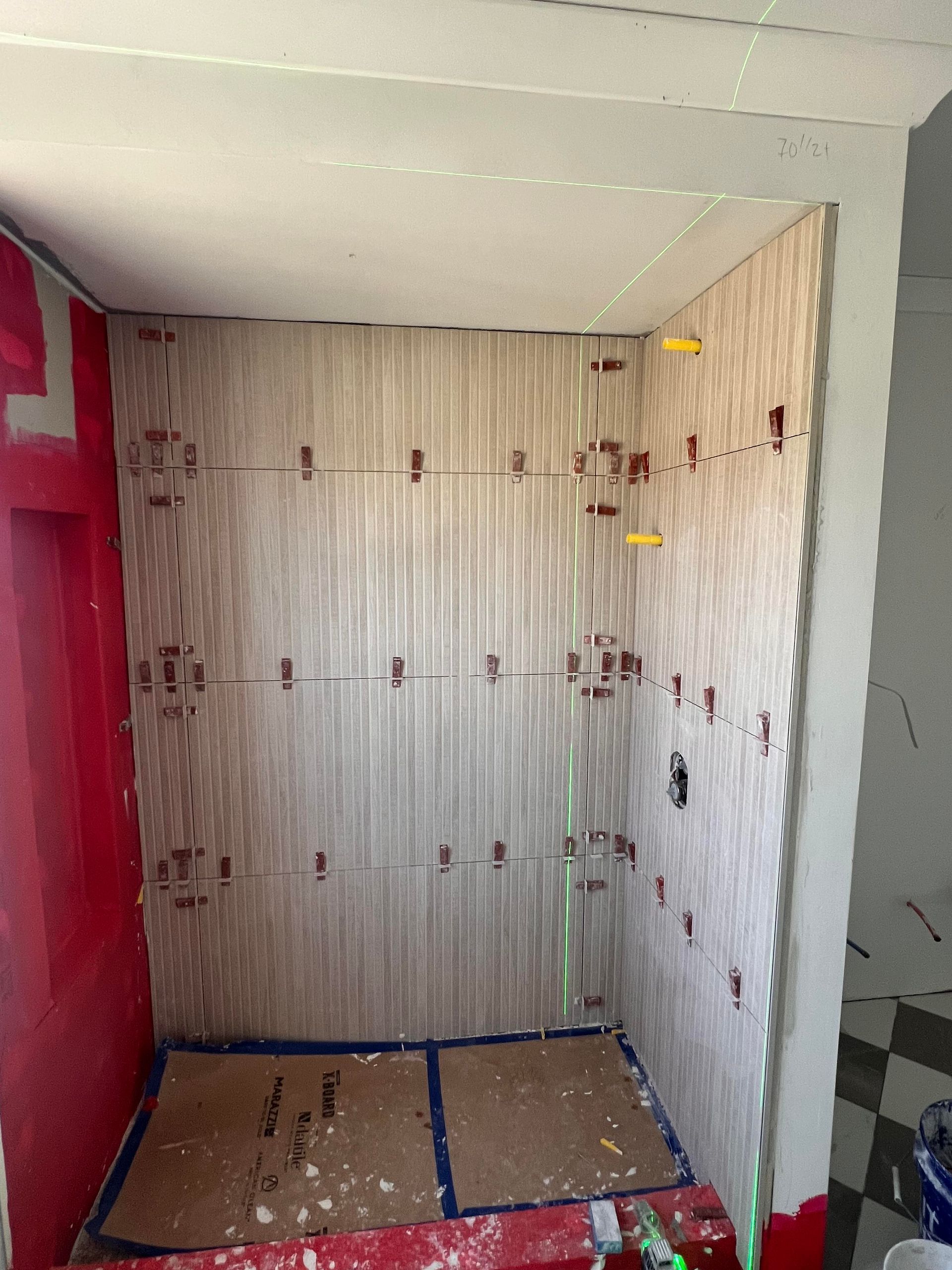 Shower stall under construction, beige tiled walls, red door frame, gray floor, spacers, and leveler.