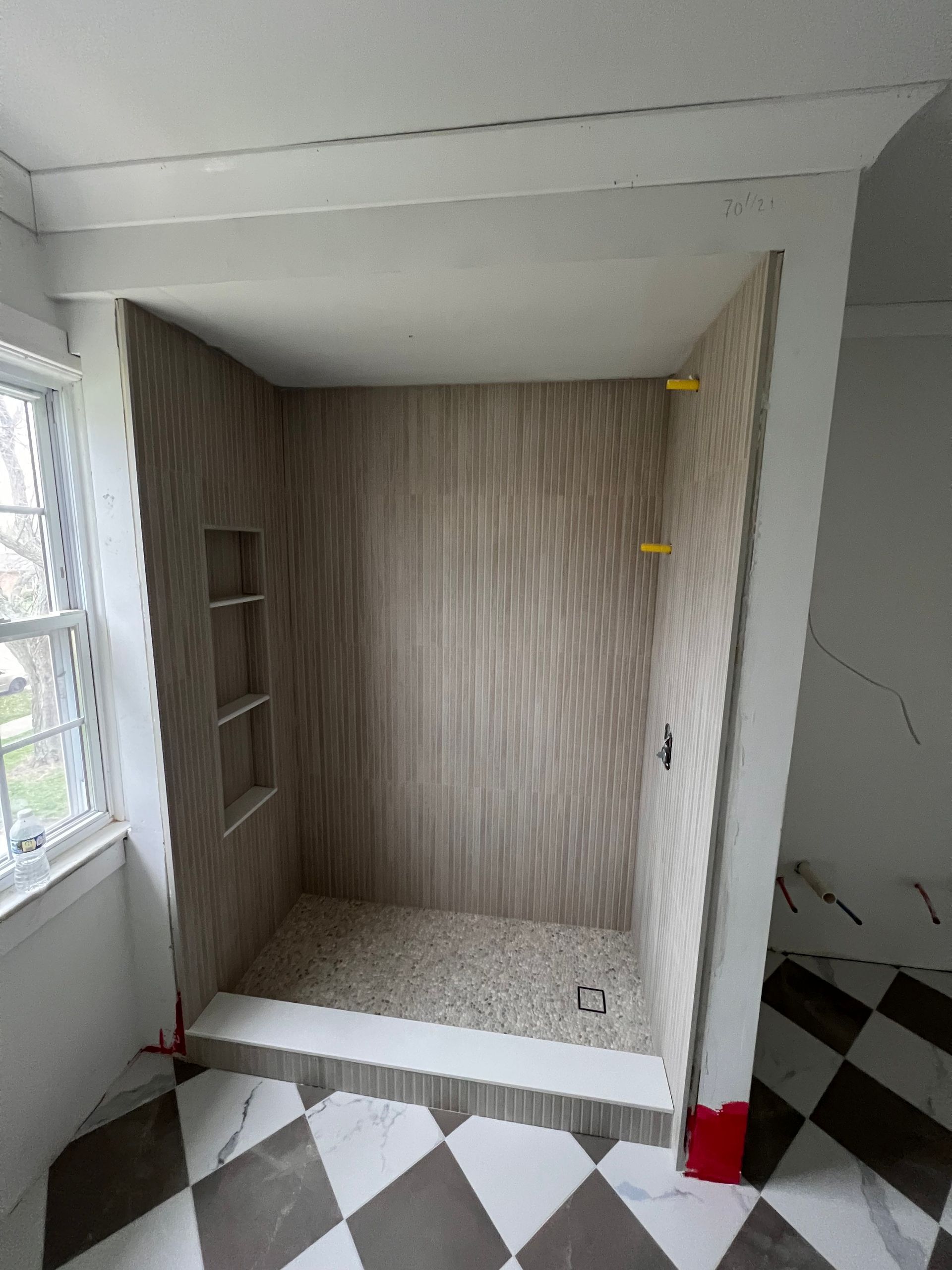 Shower stall with wood-look tile, pebble floor, and built-in shelves, in a room with checkered flooring.