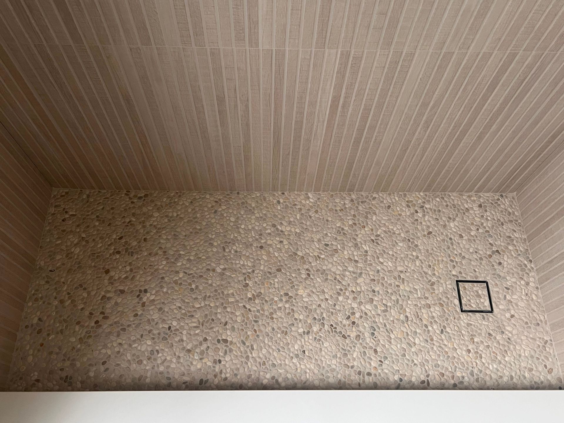 Shower stall with textured tan walls, tan slatted ceiling, and a dark square drain.
