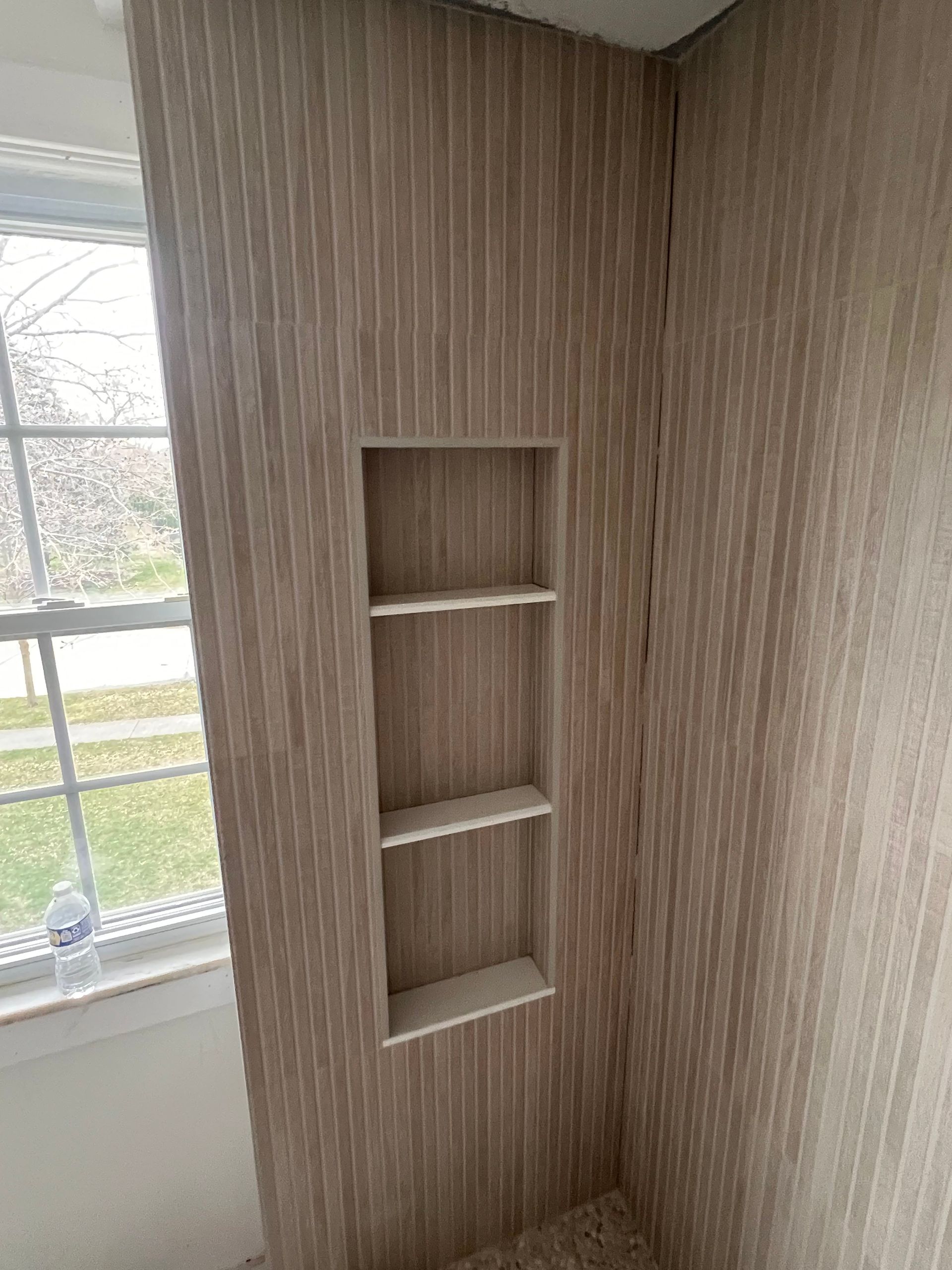 Bathroom wall with built-in shelves, window to the left, light brown vertical paneling.
