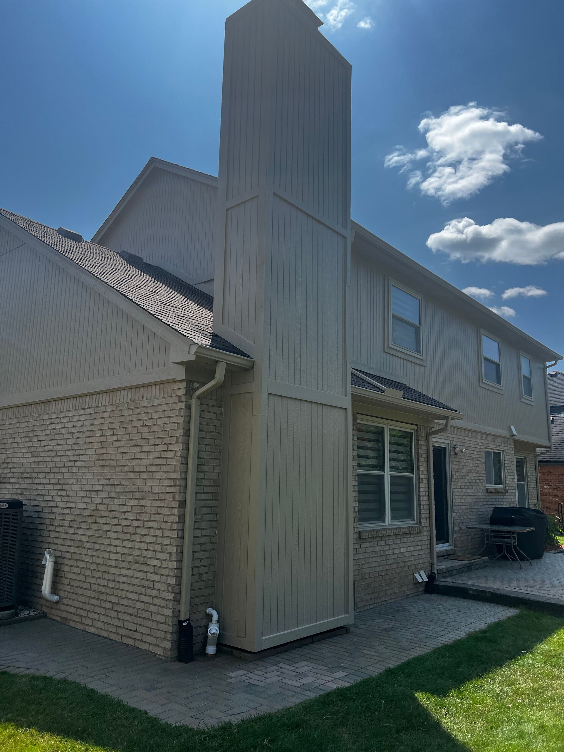 Beige brick house exterior with a chimney, a paved patio, and a grassy lawn under a blue sky.
