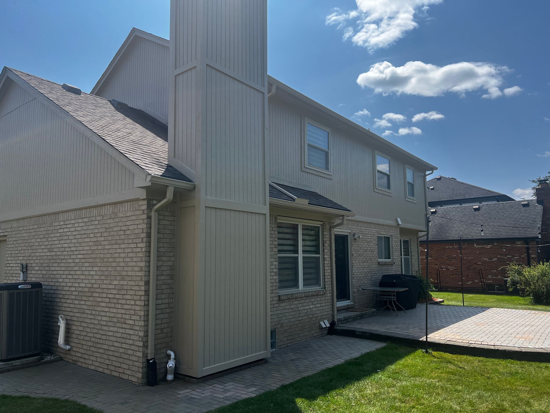 Back of a two-story beige house with a brick facade, chimney, patio, and green lawn under a sunny sky.