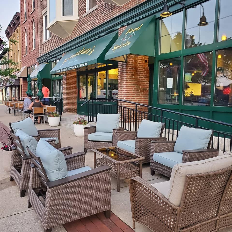 Wicker chairs and tables outside of a restaurant with a green awning that says monday