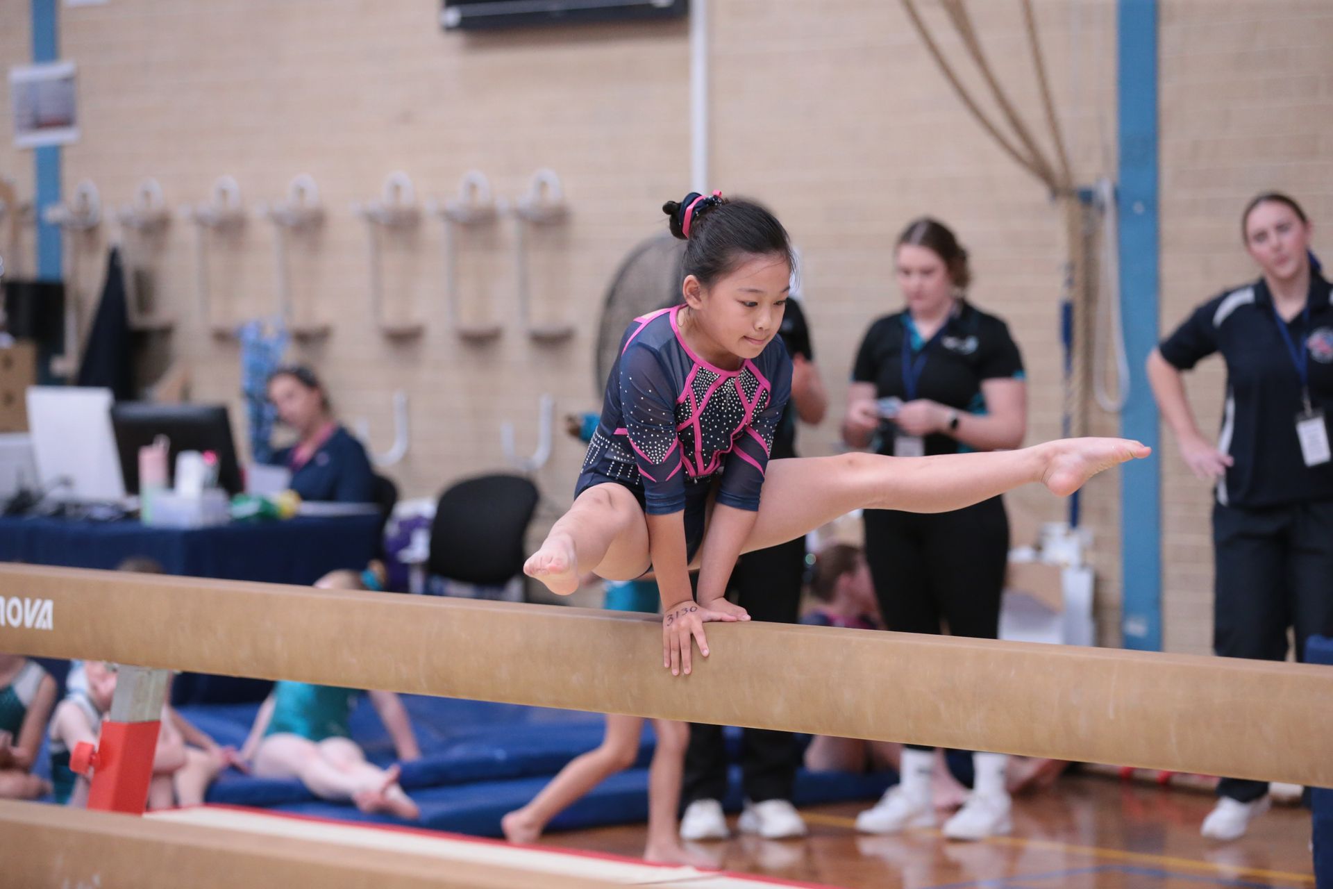 Gymnast on balance beam, performing split, wearing pink and navy leotard in competition setting.