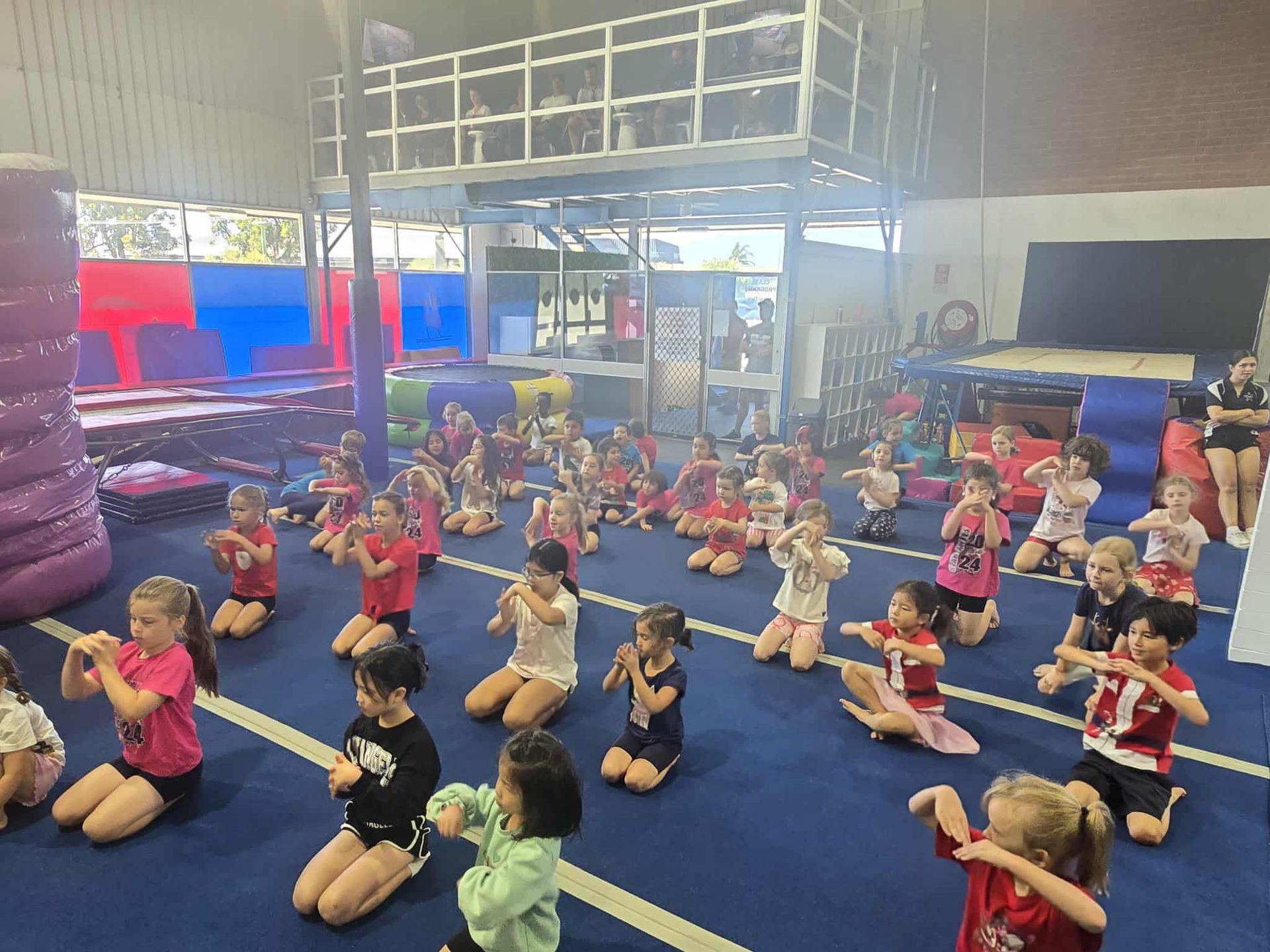 Children in red and pink shirts, kneeling on a blue mat, practicing dance moves in a gymnasium.