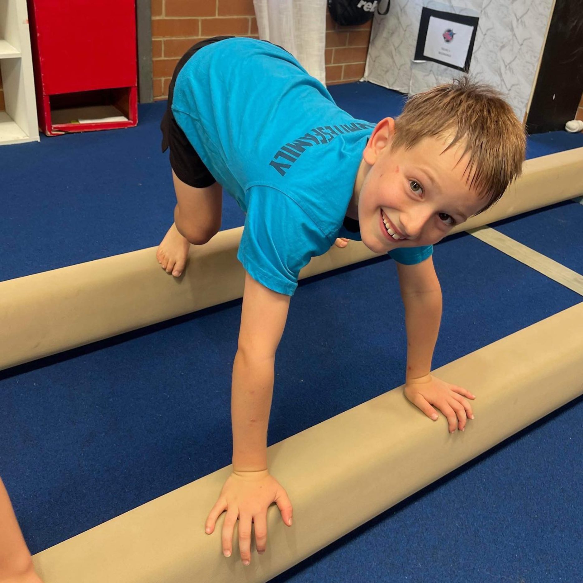 Boy smiles while crawling on balance beams in a gym; he wears blue shirt, black shorts.