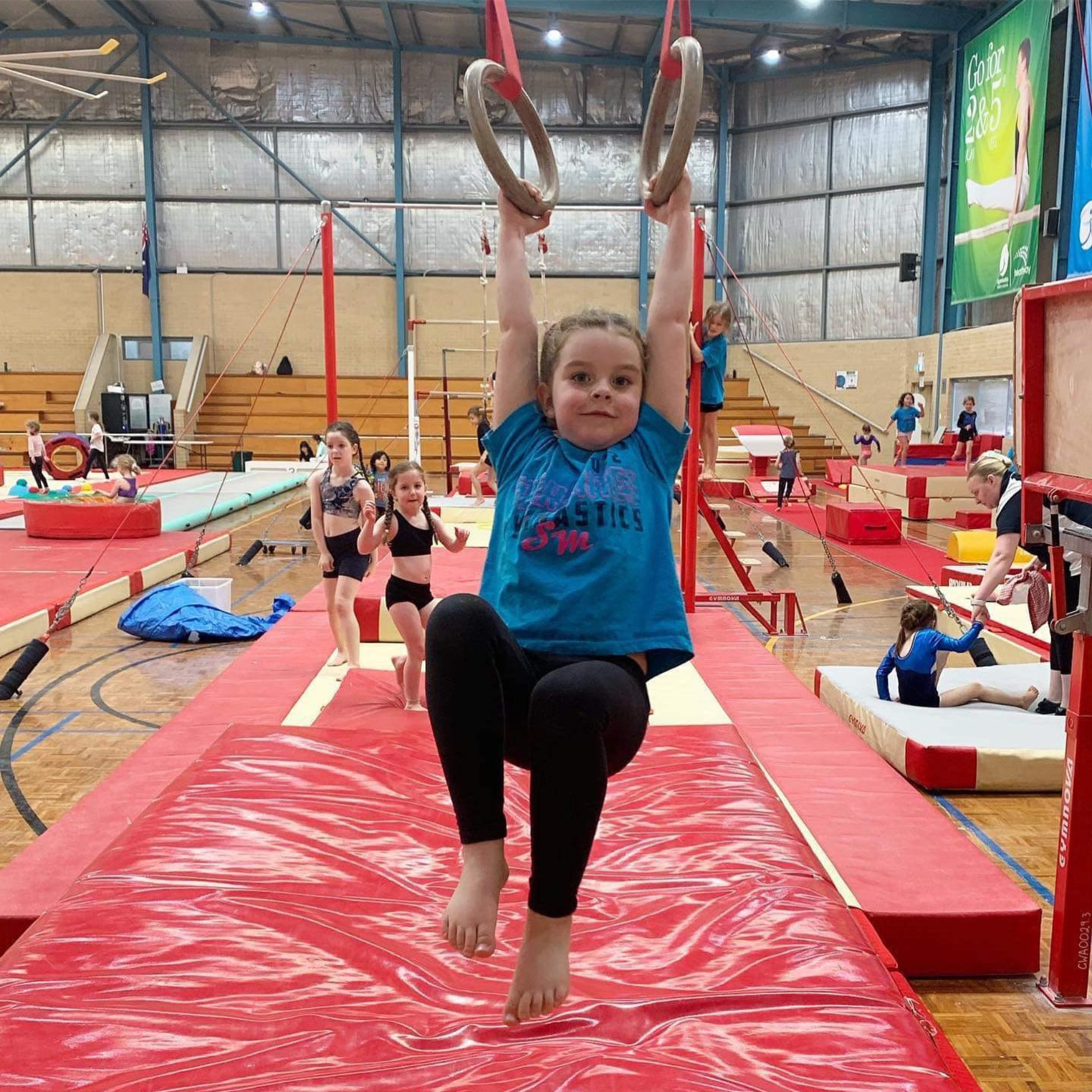 Girl on gymnastic rings in a gym, wearing blue shirt and black leggings.