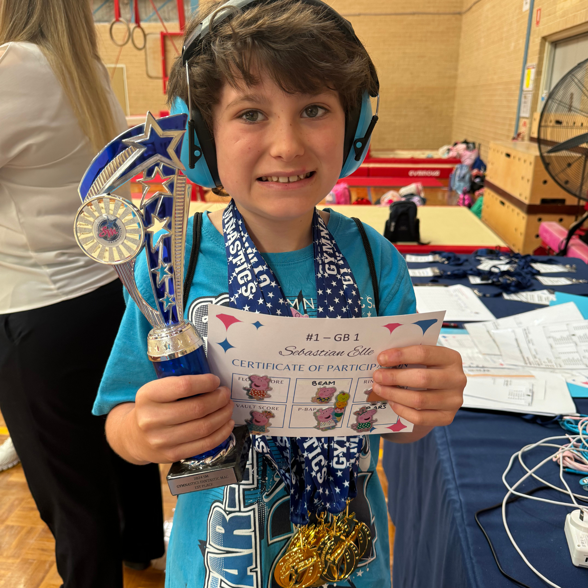 Boy wearing ear protection holds a trophy, certificate, and medals at a gymnastics event, smiling.