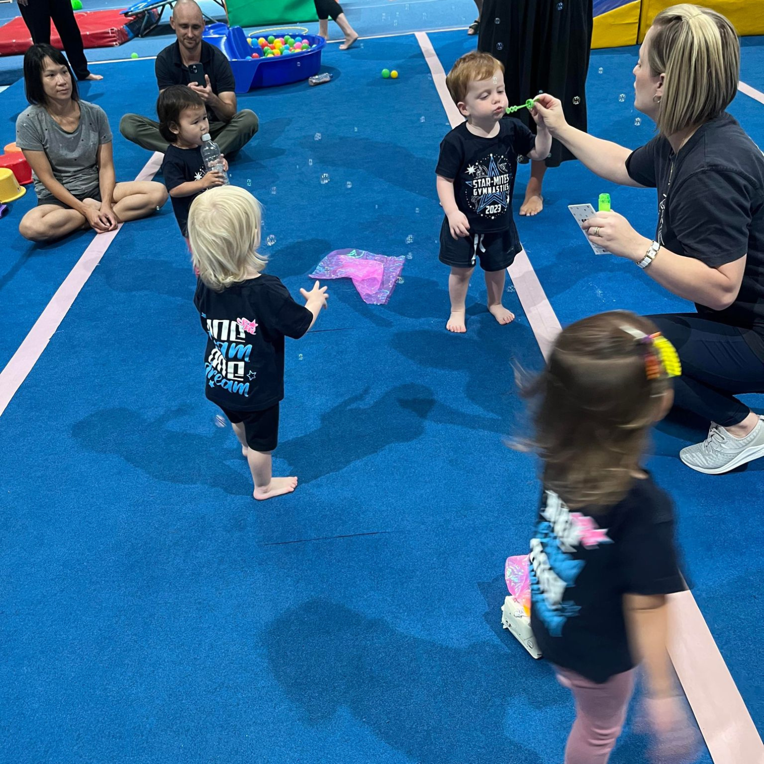 Children playing and watching bubbles in a gym. A woman blows bubbles; adults watch.