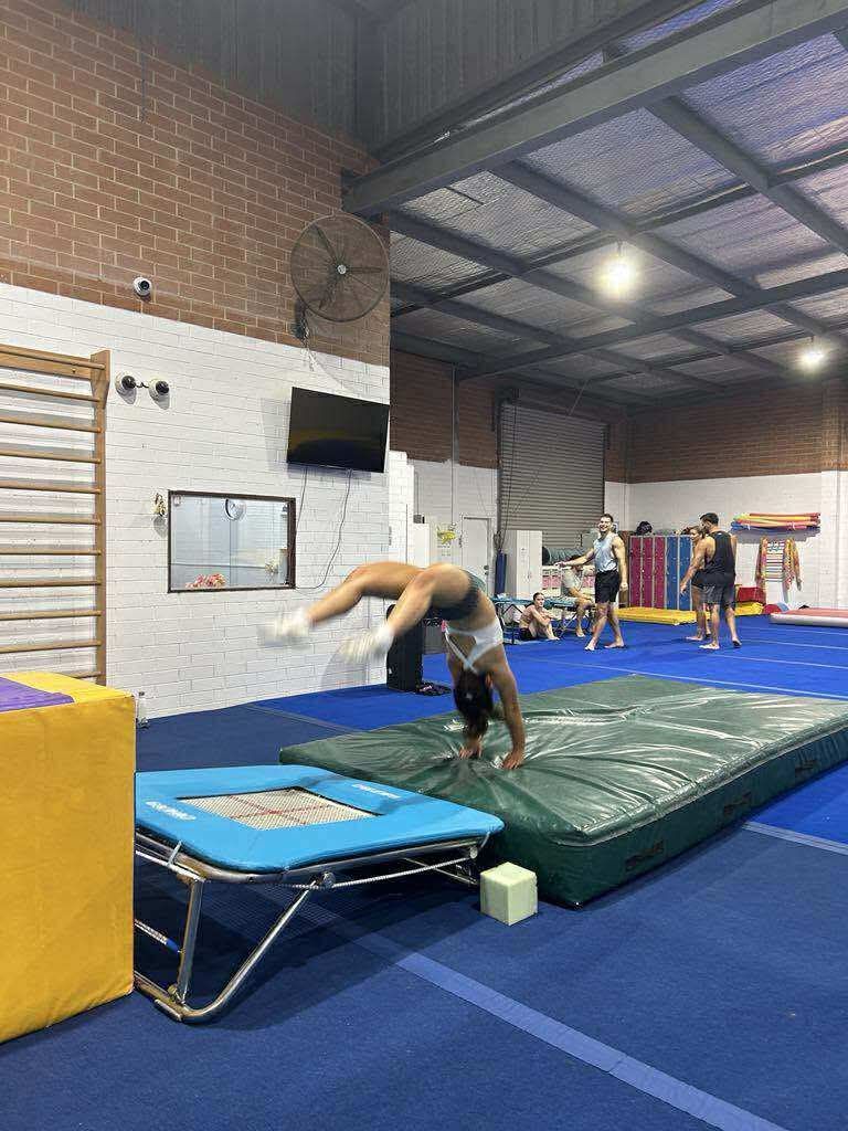 Gymnast in a backbend on a mat, near a trampoline. Gym interior with others in background.