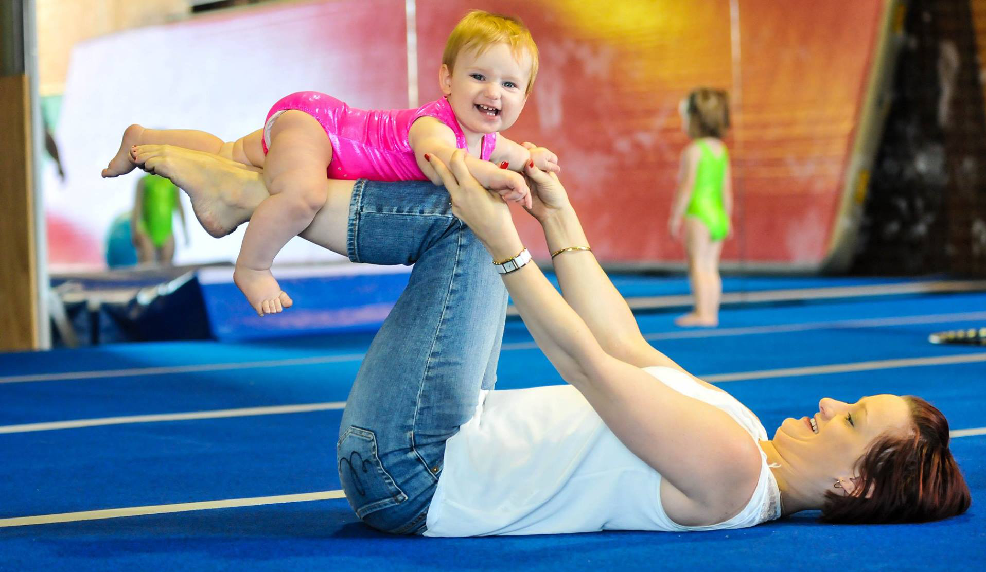 Woman lying on back with baby on feet, smiling, in a gym, blue mat.