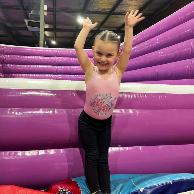 Girl in leotard and leggings with arms raised, smiling on pink and purple inflatable.
