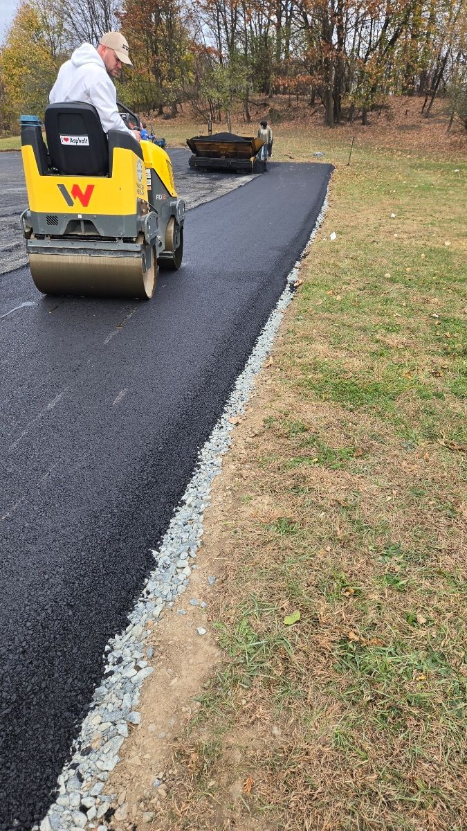 Person operating a yellow asphalt roller compacting freshly laid asphalt; a dark, new road is being created.