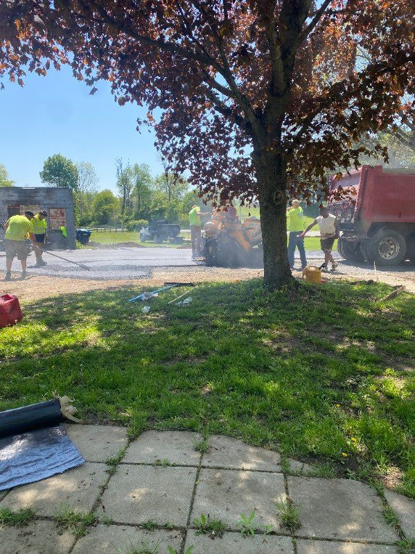 Workers paving asphalt around a tree; truck and machinery visible; sunny day.