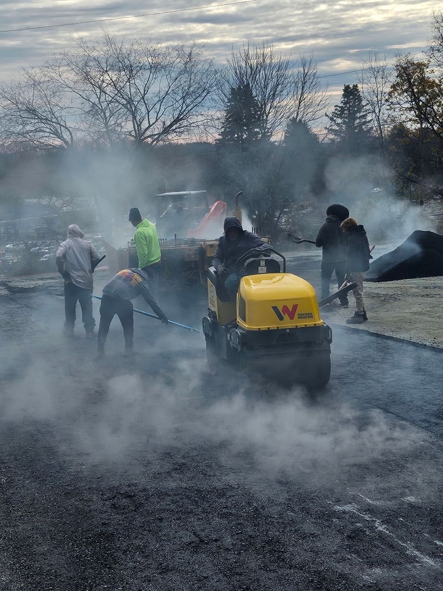 Construction workers paving asphalt with a vibratory roller. Smoke rises. Cloudy day.