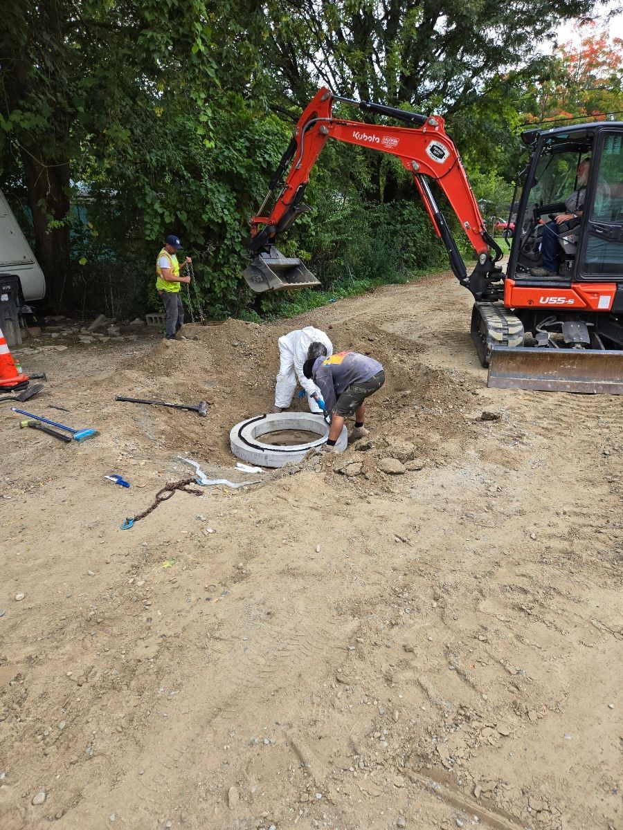 Two workers in protective gear repair a sewer access point next to a mini excavator; trees in background.