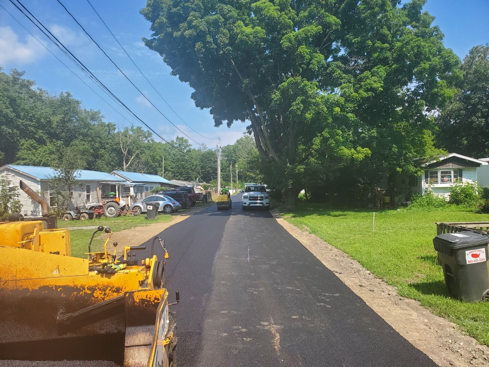 Road paving in progress. Dark asphalt covers part of street, with machinery visible. Cars and houses line the street.