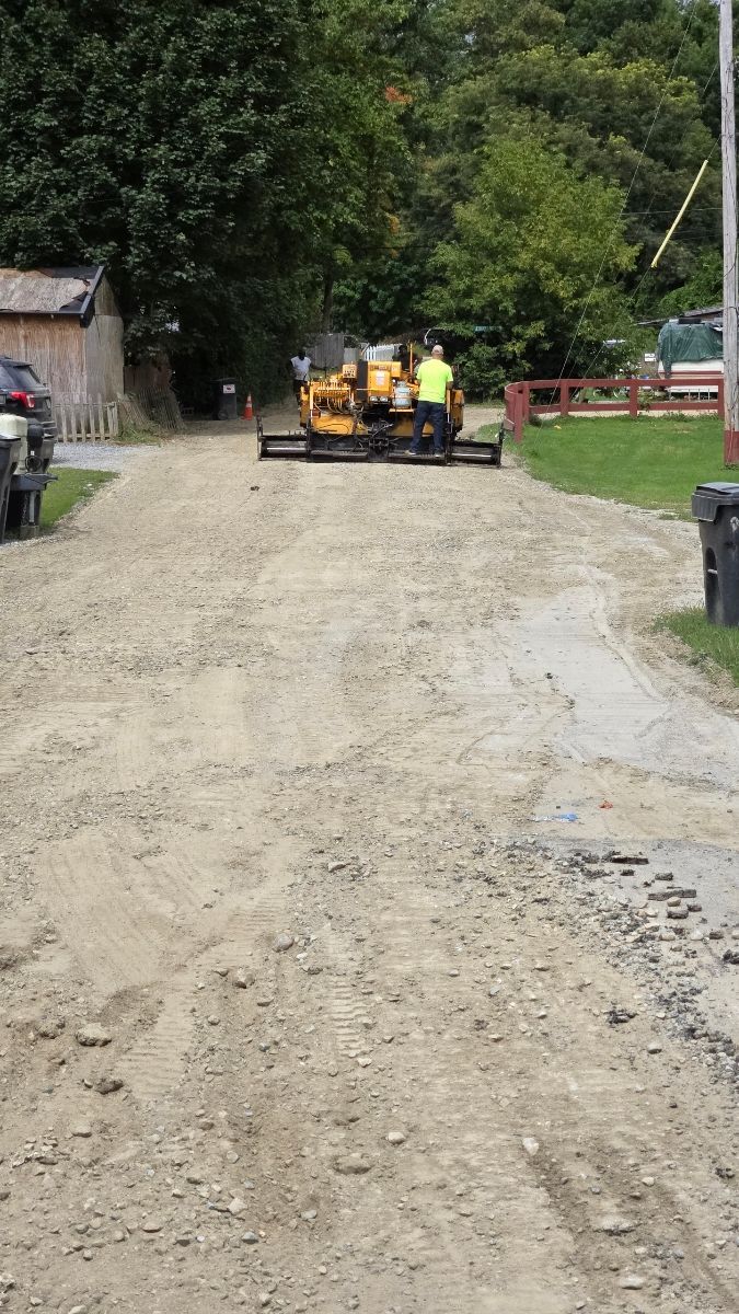 Yellow road grader smoothing a gravel road. A worker stands nearby.