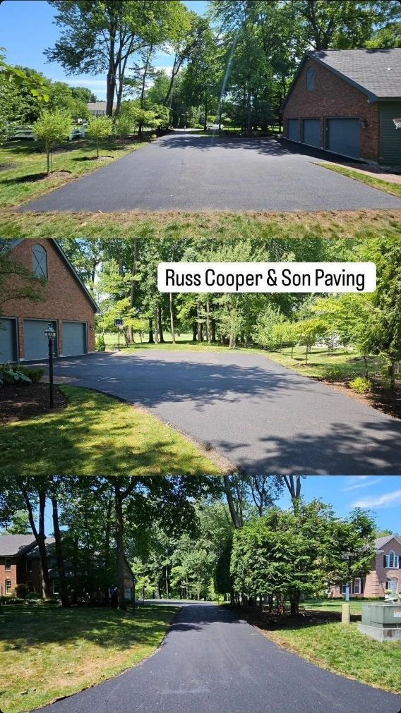 Black asphalt driveway leading to a brick garage, surrounded by green trees and grass.