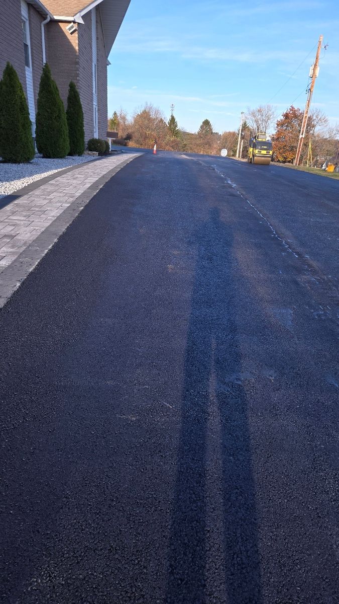 Asphalt road, person's shadow cast in the center. Building and trees on the left; blue sky background.