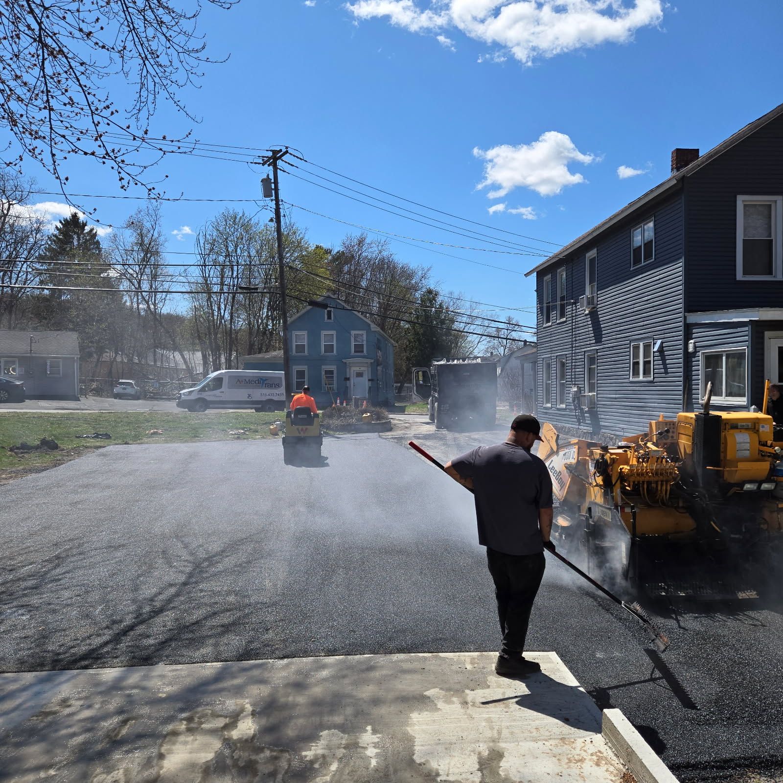 Asphalt paving in progress on a sunny day. Worker raking hot asphalt. Roller compacting the freshly paved surface. Buildings and sky in background.