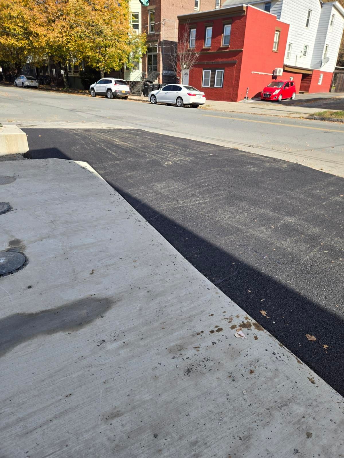 Newly paved asphalt next to a concrete sidewalk on a city street. Cars parked on the street, red building in background.