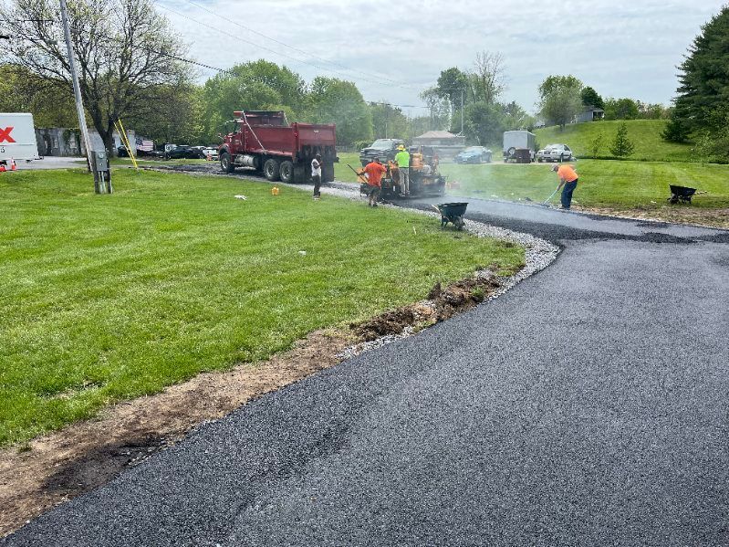 Asphalt paving in progress: workers, truck, paving machine, and dark asphalt on grass next to a road, overcast sky.
