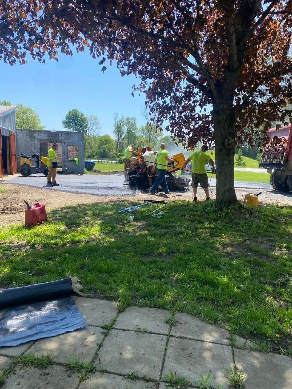 Asphalt paving crew working outdoors, using machinery and wearing yellow vests near a tree.