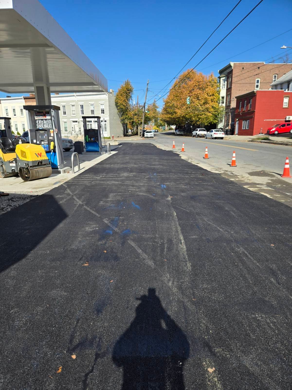 Freshly paved road beside a gas station with a blue sky, autumn trees, and orange traffic cones.