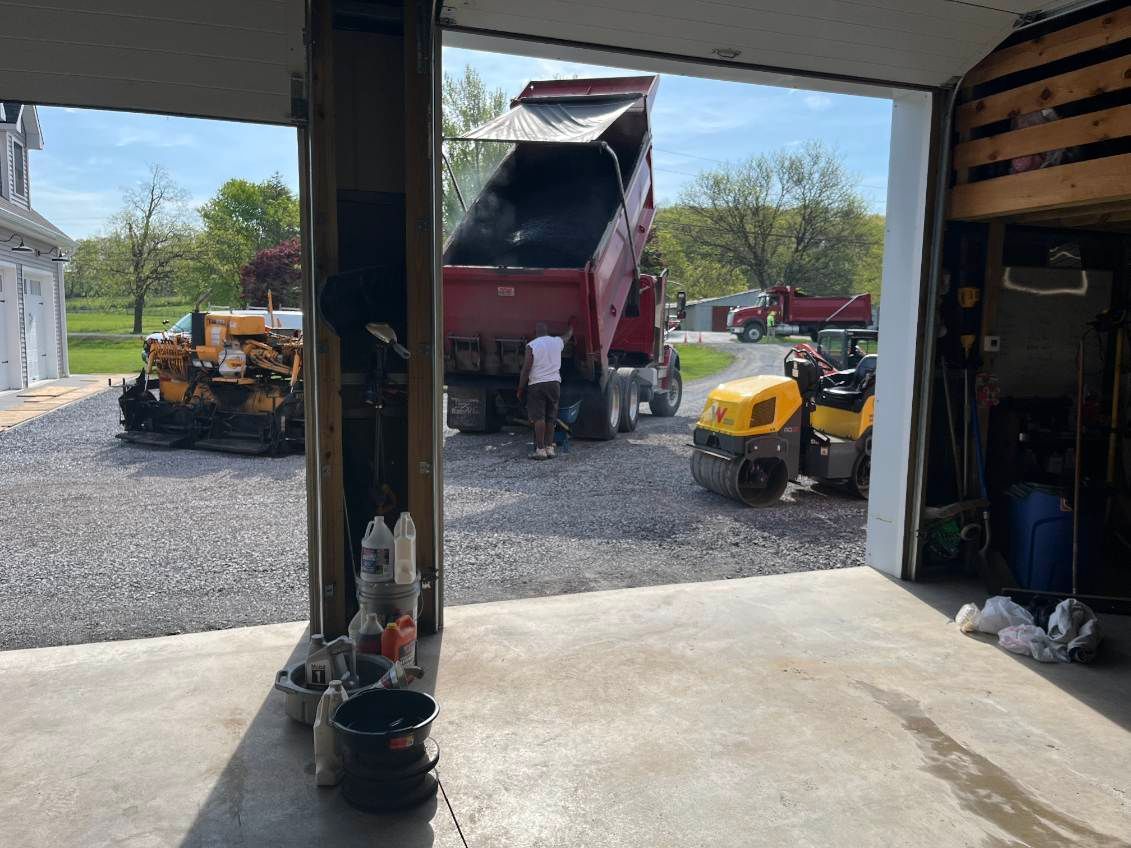 A truck dumping asphalt onto a gravel driveway; construction equipment nearby, seen from a garage.