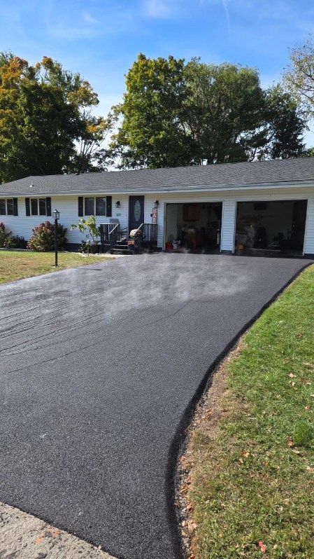 Black paved driveway leading to a white house with a two-car garage under a blue sky.
