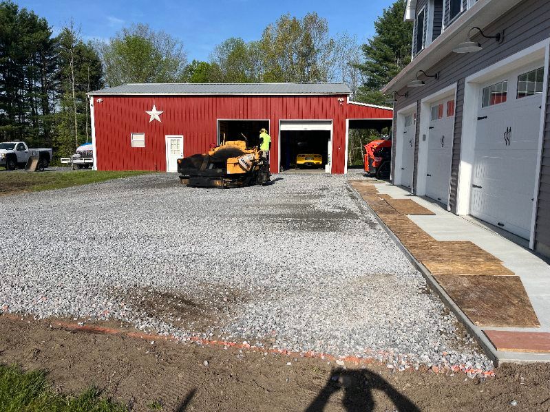 Gravel driveway construction with red barn and garage. Construction equipment, a worker, and a concrete ramp are visible.