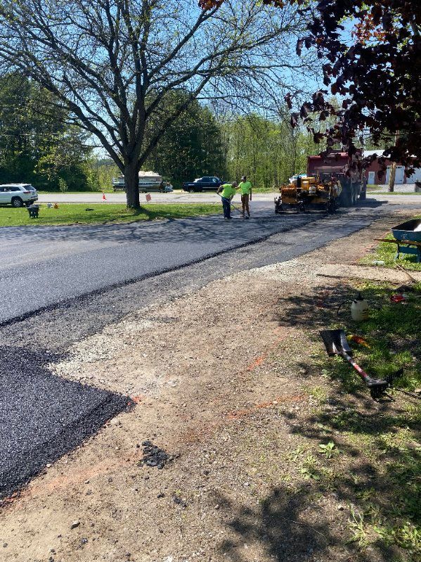 Road paving in progress; workers in neon vests, asphalt machine, fresh blacktop, gravel shoulder, sunny day.