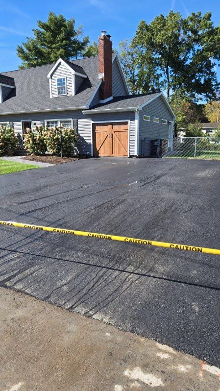Newly paved asphalt driveway with yellow caution tape, leading to a gray house with a brown garage door.