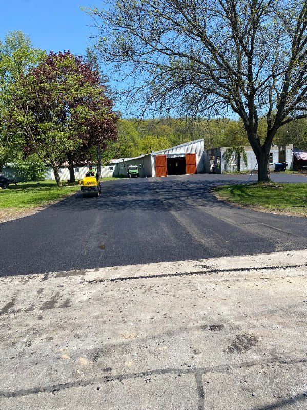 Freshly paved asphalt driveway leading to a white building with orange doors; tree on right.