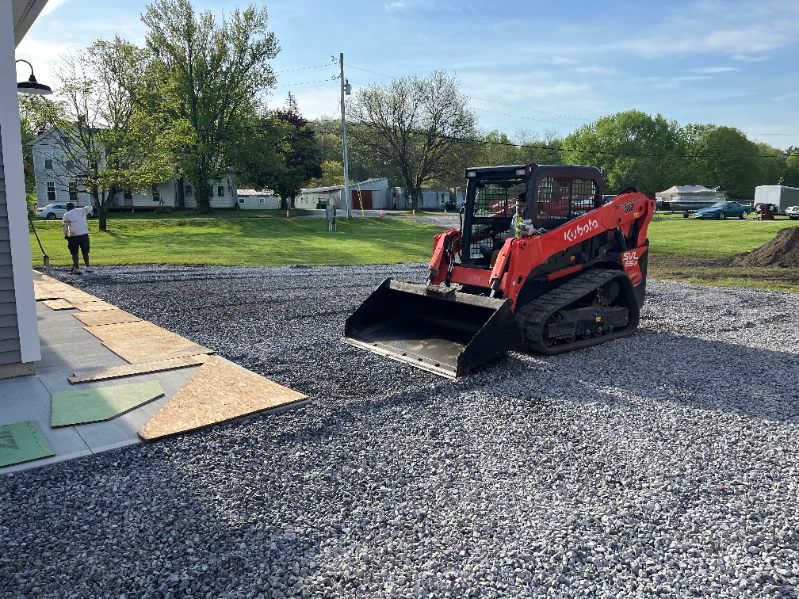 A red Kubota skid steer on gravel, next to a building. A person rakes gravel nearby. Outdoors.