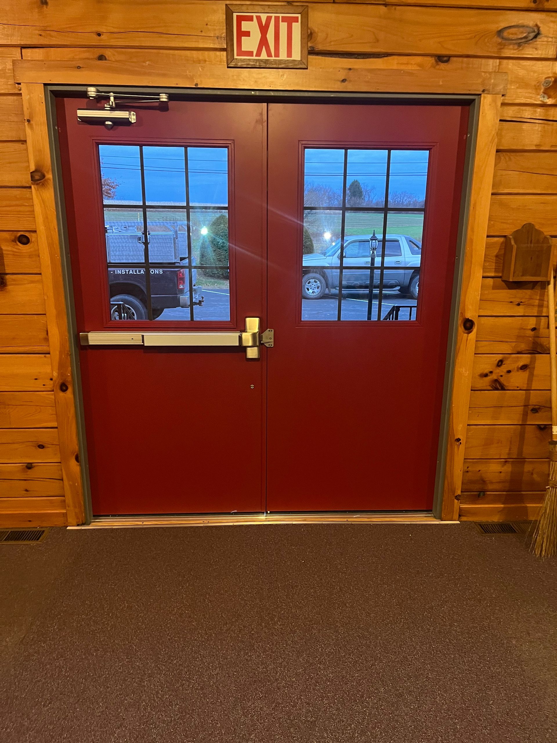 A pair of red double exit doors with windows, set in a log cabin wall with an illuminated “EXIT” sign above.