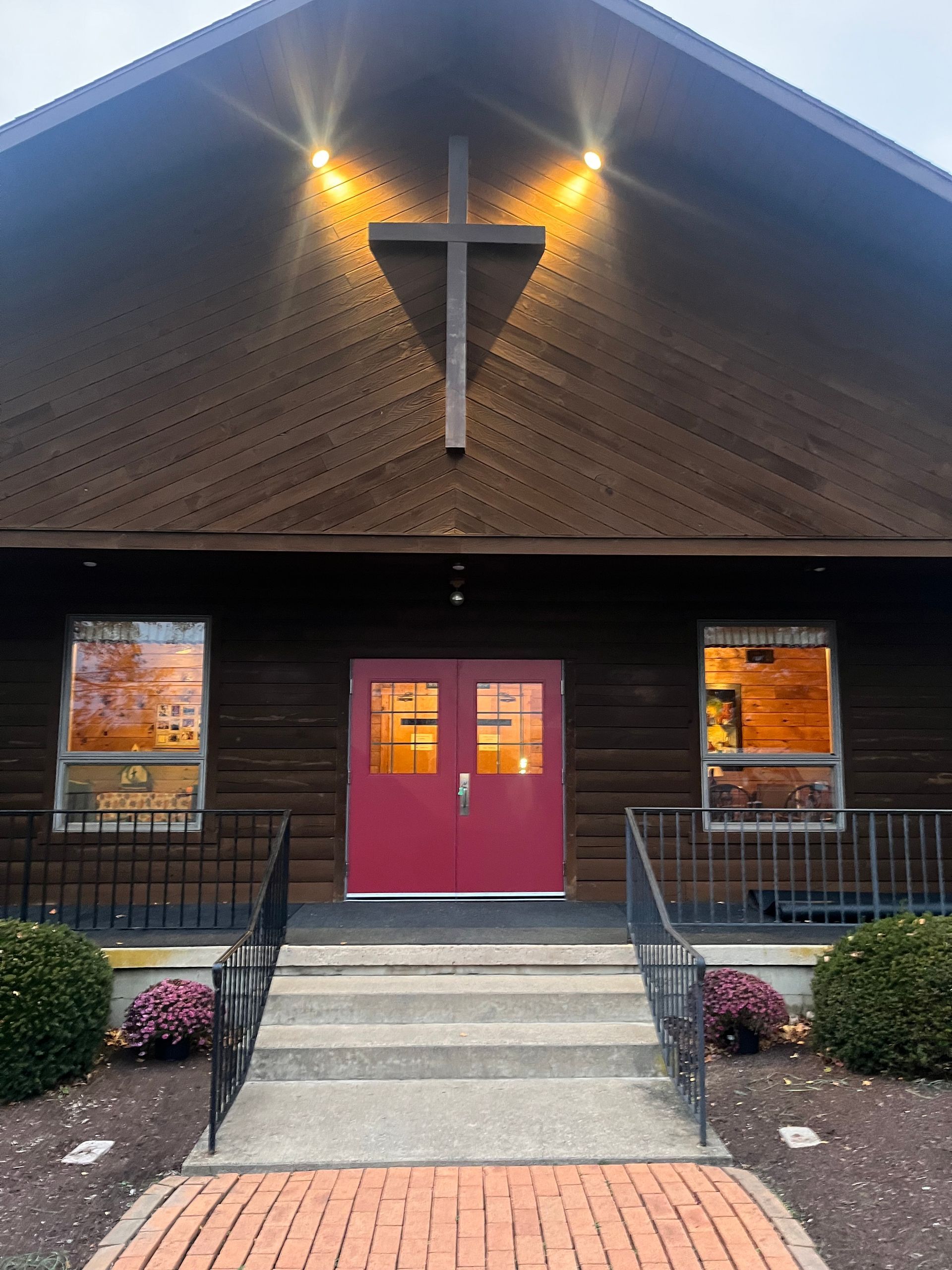 A wood-paneled building with a central cross, lit by two exterior lights, featuring double red doors and concrete steps.