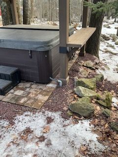 Outdoor hot tub and wooden deck beside a snowy forest path with rocks and tree trunk