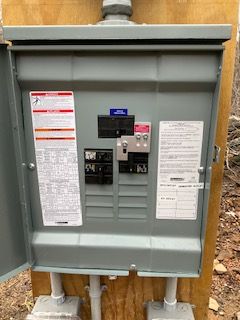 Gray electrical meter and breaker panel mounted outdoors on plywood, with warning labels and conduit pipes.