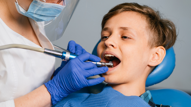 Dentist treating a child’s teeth with a drill while the child sits in a blue chair