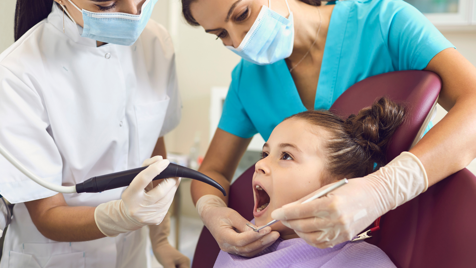 Dentist treating a young patient while a nurse assists in a clinic, all wearing masks and gloves