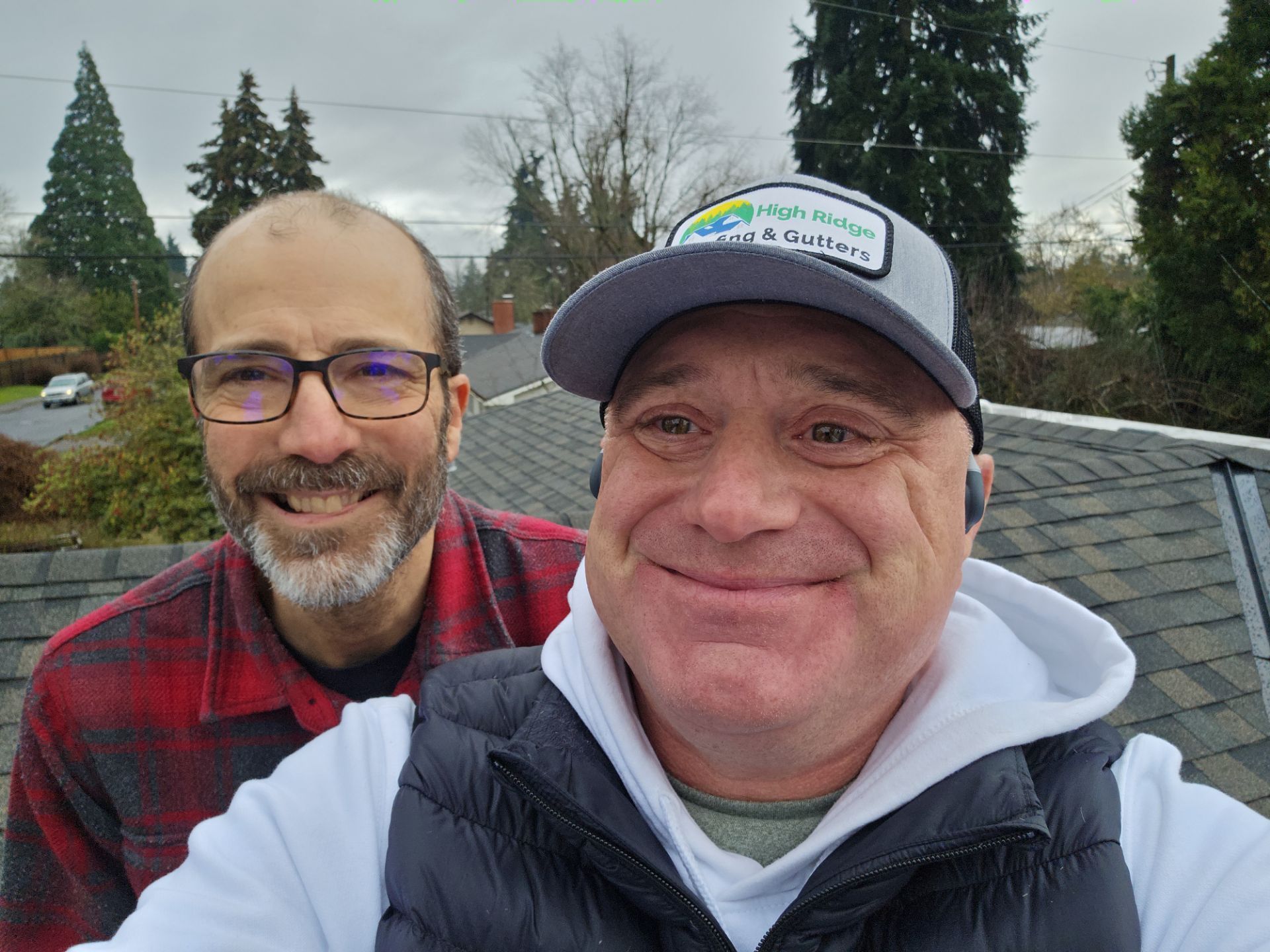 Two smiling men posing outdoors on a cloudy day, one in a red plaid shirt and one in a cap and hoodie.
