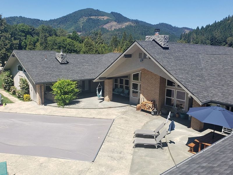 Mountain lodge with gray shingle roof and patio, set against forested hills under a clear sky