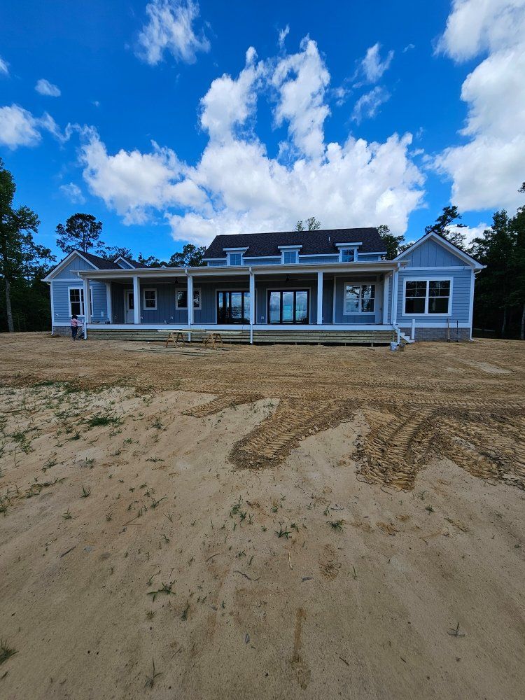 A light blue single-story house with a wrap-around porch under construction, situated on a large, sandy, cleared lot.
